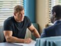 Actor Alan Ritchson sits at a table in a muscle-bound t-shirt during a scene from Amazon's