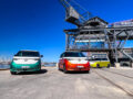a green and white ID Buzz, an orange and white ID Buzz, and a yellow and white VW ID Buzz parked underneath a dock crane