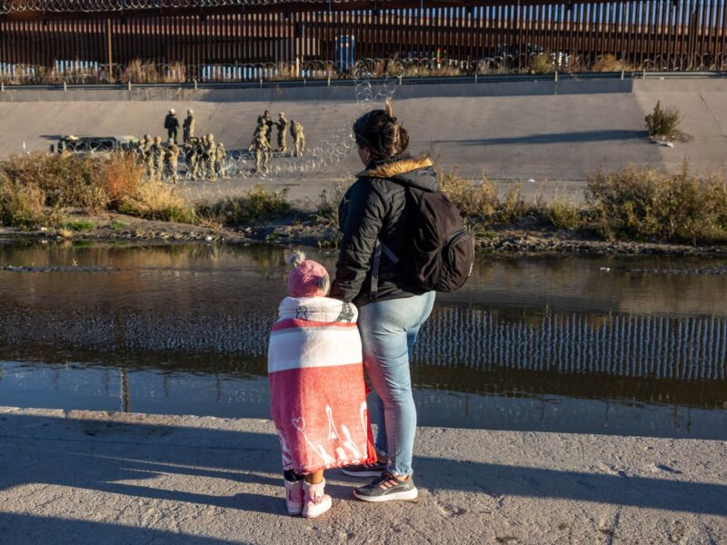 A woman and child look at uniformed men behind loops of concertina wire across a river channel