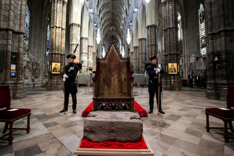 A big hunk of rectangular rock on a platform in front of a  throne, flanked by two men in uniform.