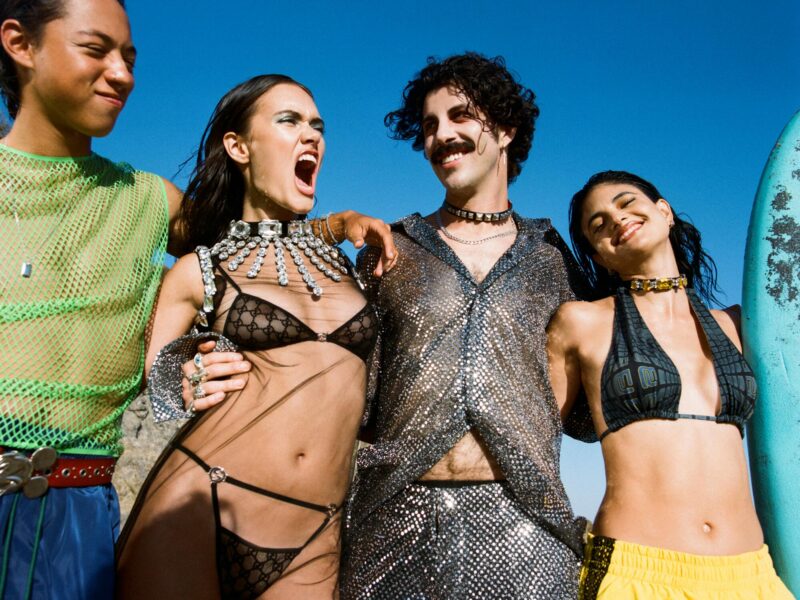 Four people wearing colorful outfits on a beach embrace.