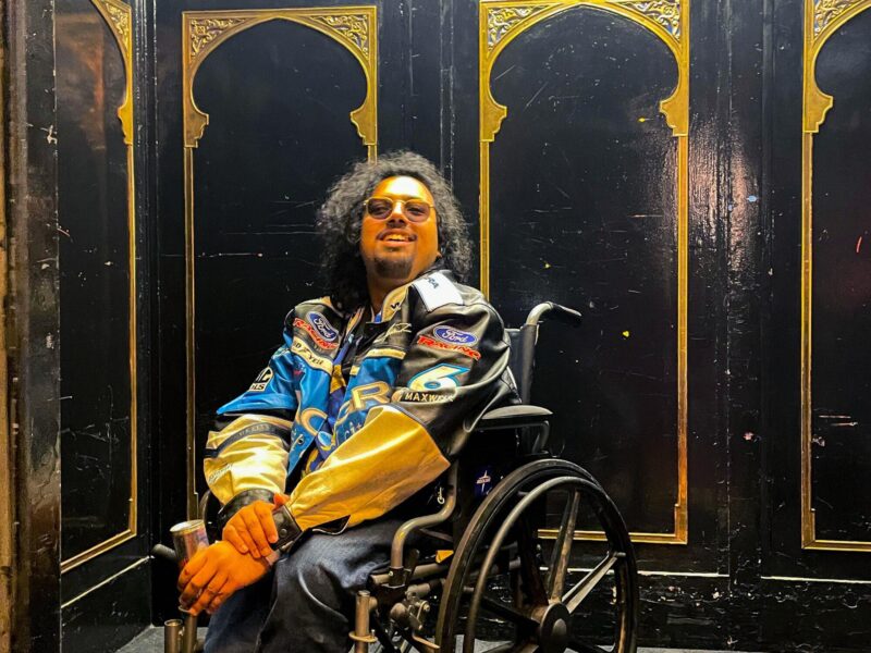 Saptarshi Majumdar wearing a racing jacket sits in a wheelchair in an elevator with black walls and gold trim.